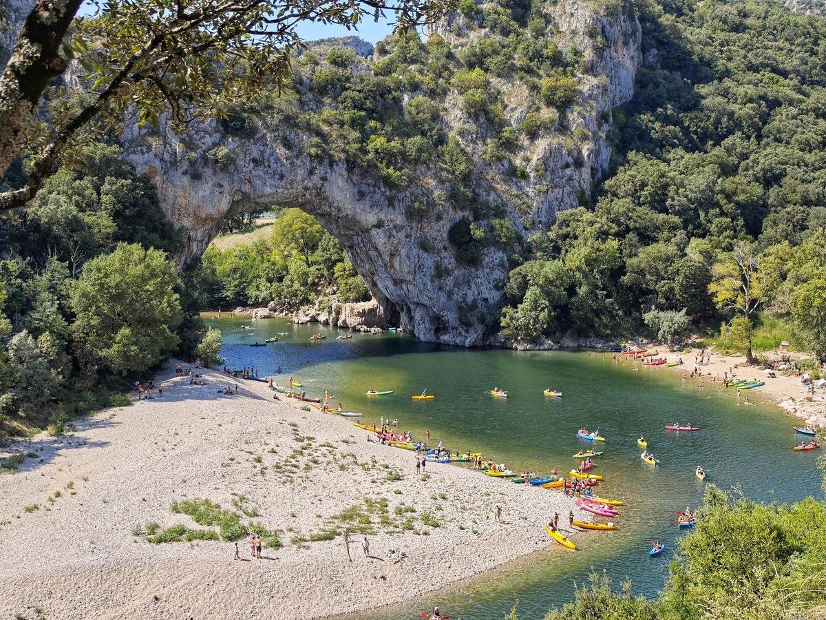 Vue du Pont d’Arc en Ardèche avec de nombreux canoës colorés sur la rivière et des vacanciers profitant de la plage.
