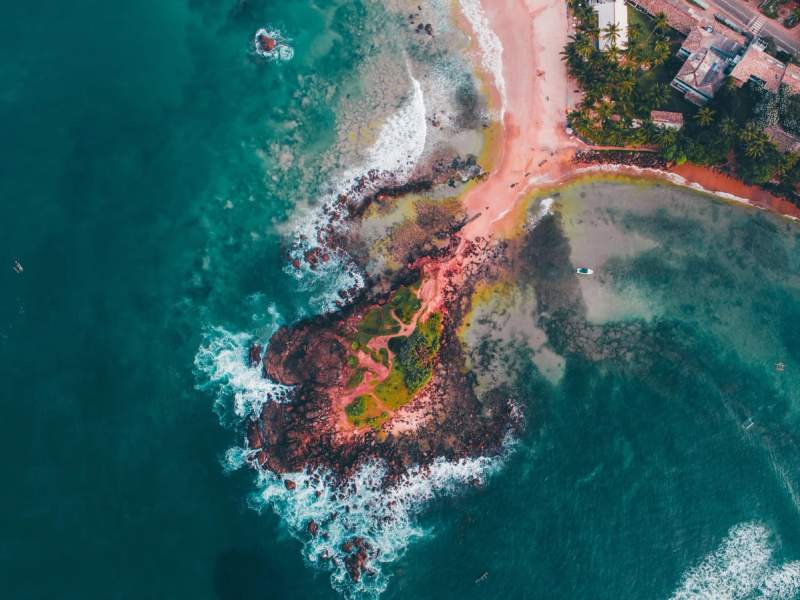 Vue aérienne de Parrot Rock à Mirissa, Sri Lanka, montrant un îlot rocheux entouré d'eaux turquoise avec des vagues s'écrasant contre les rochers. Une fine bande de sable relie l'îlot à la plage, bordée de palmiers et de bâtiments en arrière-plan.