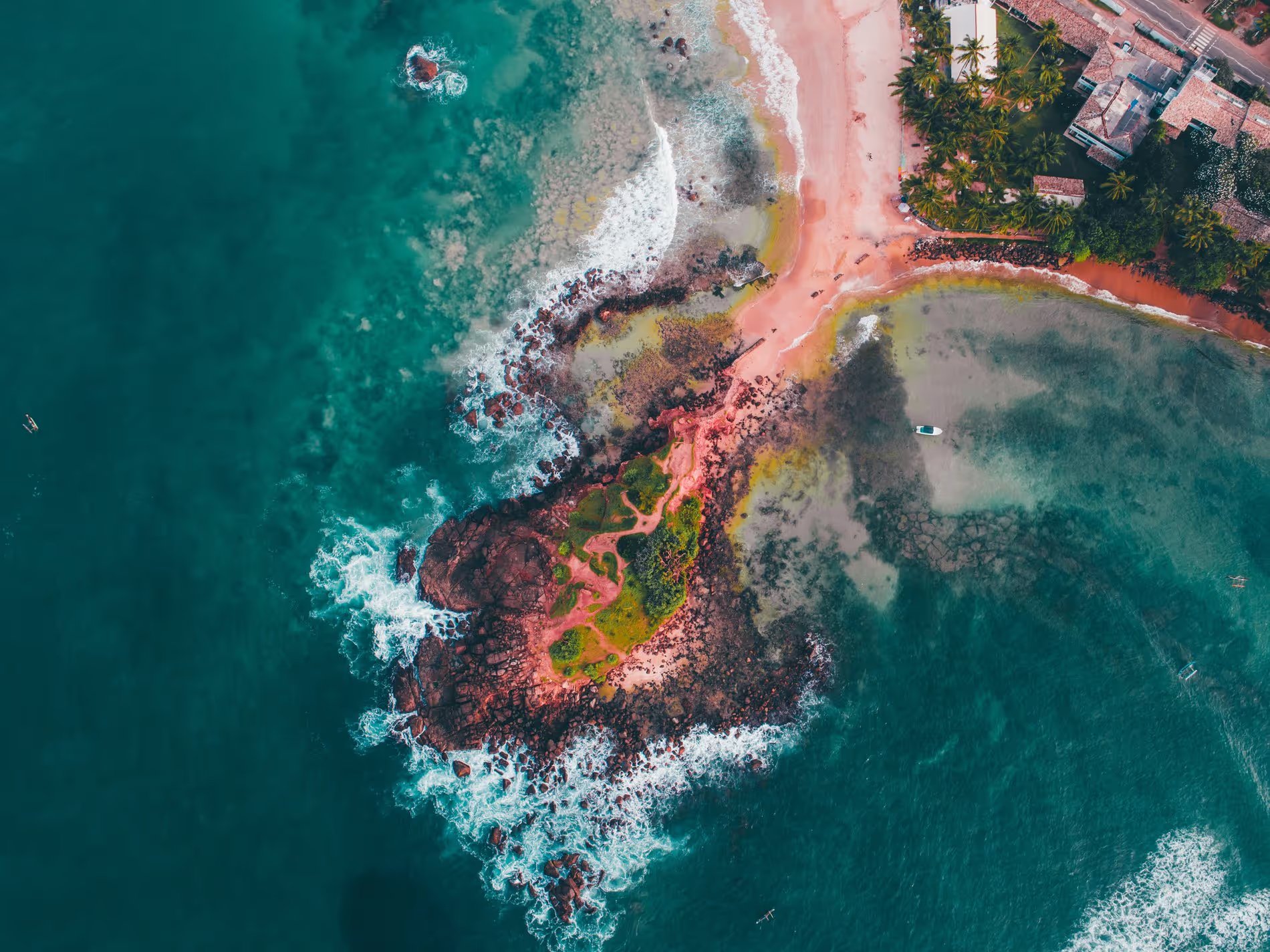 Vue aérienne de Parrot Rock à Mirissa, Sri Lanka, montrant un îlot rocheux entouré d'eaux turquoise avec des vagues s'écrasant contre les rochers. Une fine bande de sable relie l'îlot à la plage, bordée de palmiers et de bâtiments en arrière-plan.