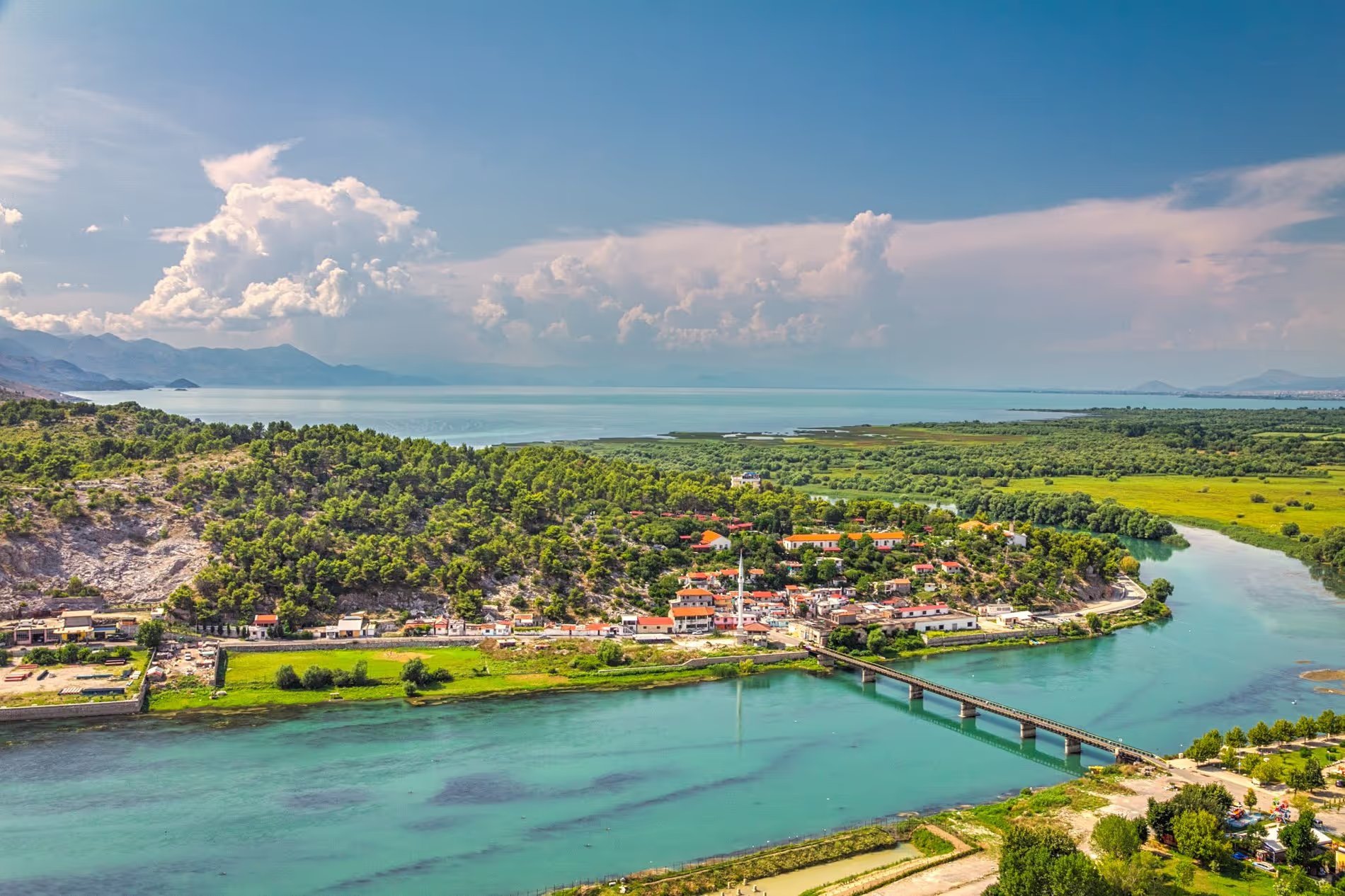 Vue panoramique sur la ville de Shkodër avec la rivière, les collines et le lac Skadar en arrière-plan sous un ciel dégagé.