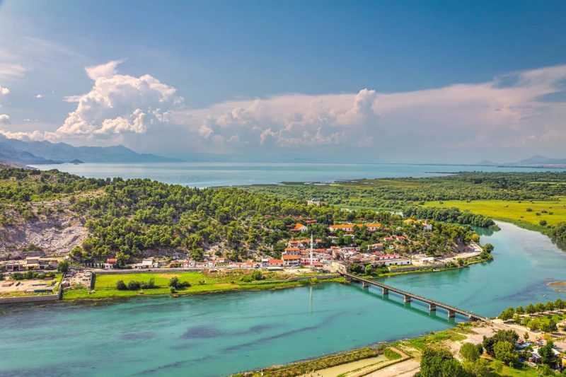 Vue panoramique sur la ville de Shkodër avec la rivière, les collines et le lac Skadar en arrière-plan sous un ciel dégagé.