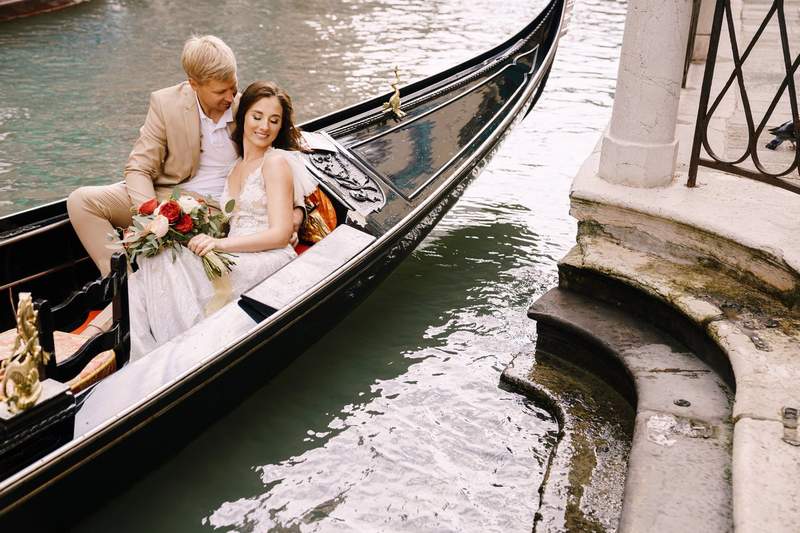 Couple de mariés enlacés dans une gondole sur un canal de Venise, la mariée tenant un bouquet de roses rouges et blanches