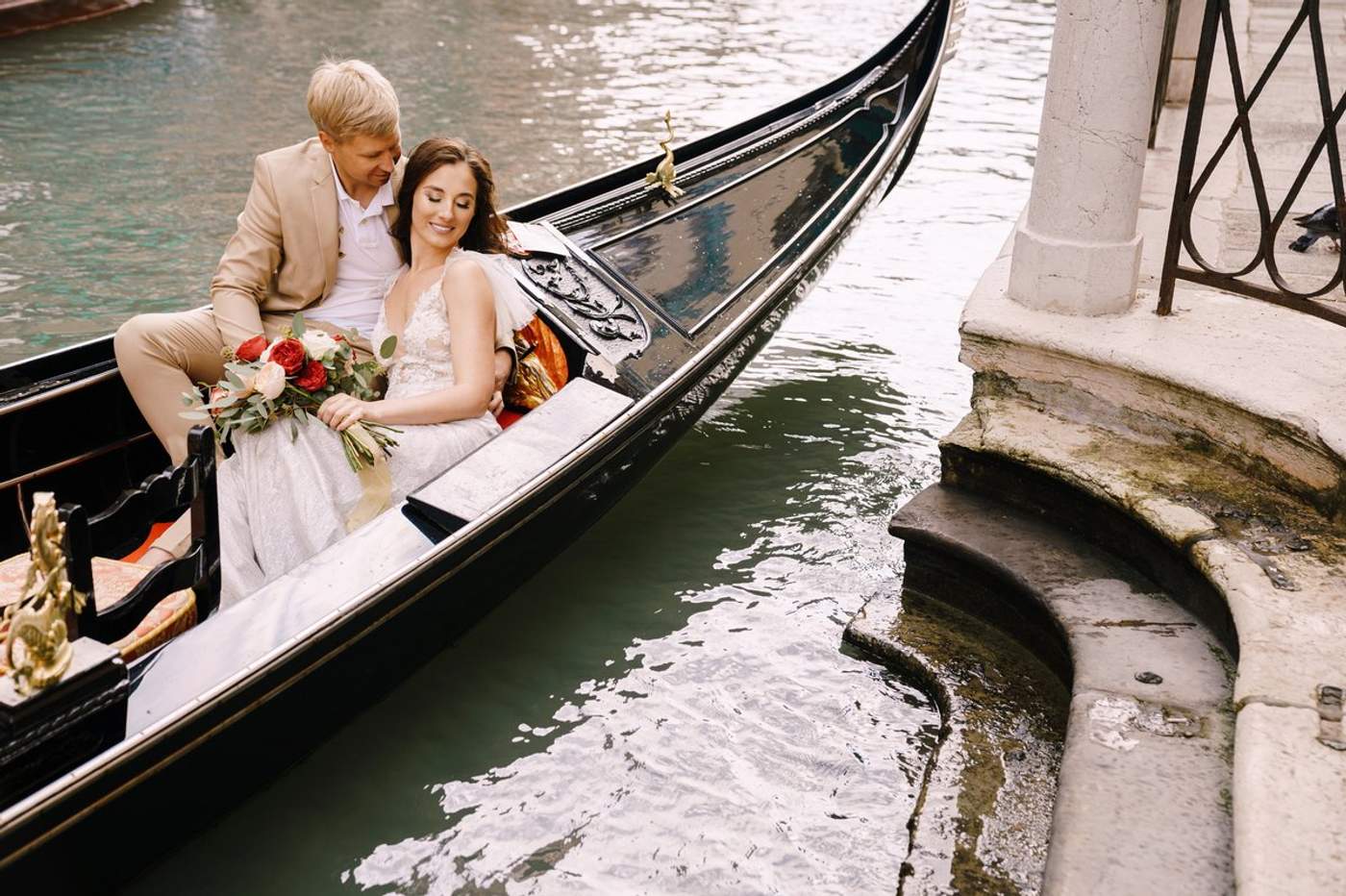 Couple de mariés enlacés dans une gondole sur un canal de Venise, la mariée tenant un bouquet de roses rouges et blanches