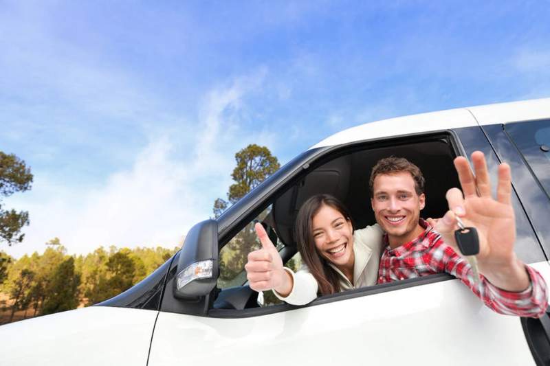 Couple souriant à l’intérieur d’un véhicule blanc, tenant une clé de voiture et faisant un geste de réussite, avec un ciel bleu en arrière-plan.