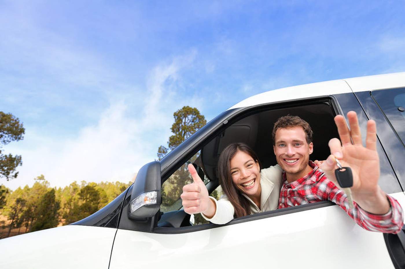 Couple souriant à l’intérieur d’un véhicule blanc, tenant une clé de voiture et faisant un geste de réussite, avec un ciel bleu en arrière-plan.