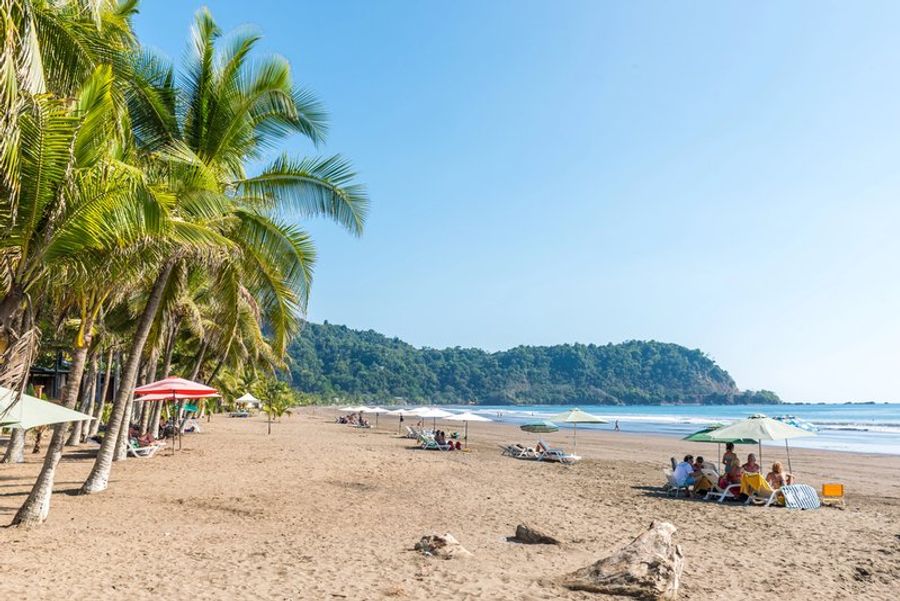 Plage bordée de palmiers à Jacó avec des visiteurs profitant du soleil.