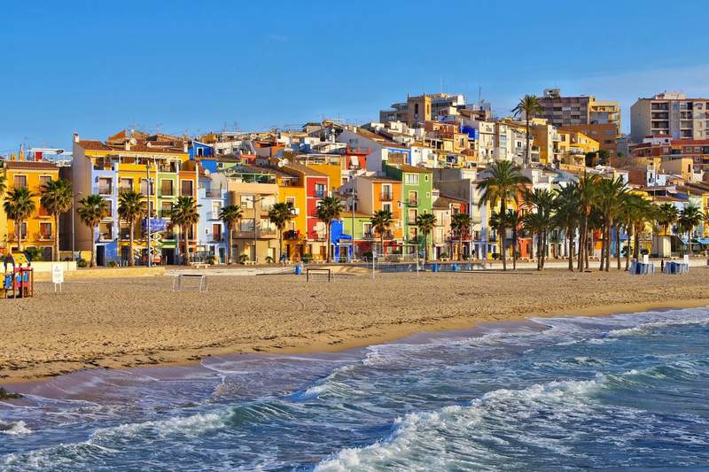 Maisons colorées de Villajoyosa sur la Costa Blanca, Espagne, avec la plage et les palmiers au premier plan sous un ciel bleu lumineux.