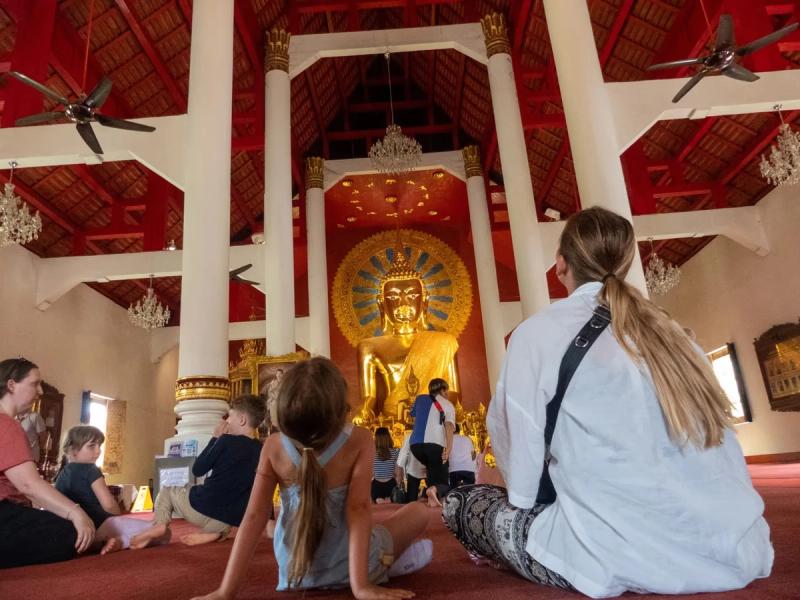 Intérieur d'un temple à Chian Mai en Thaïlande avec une grande statue dorée de Bouddha, des colonnes blanches et des visiteurs assis sur le sol, regardant la statue.