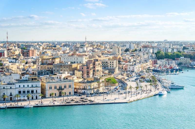Vue panoramique du port de Brindisi en Italie, avec ses bâtiments historiques, sa promenade animée et ses bateaux amarrés dans une eau turquoise.