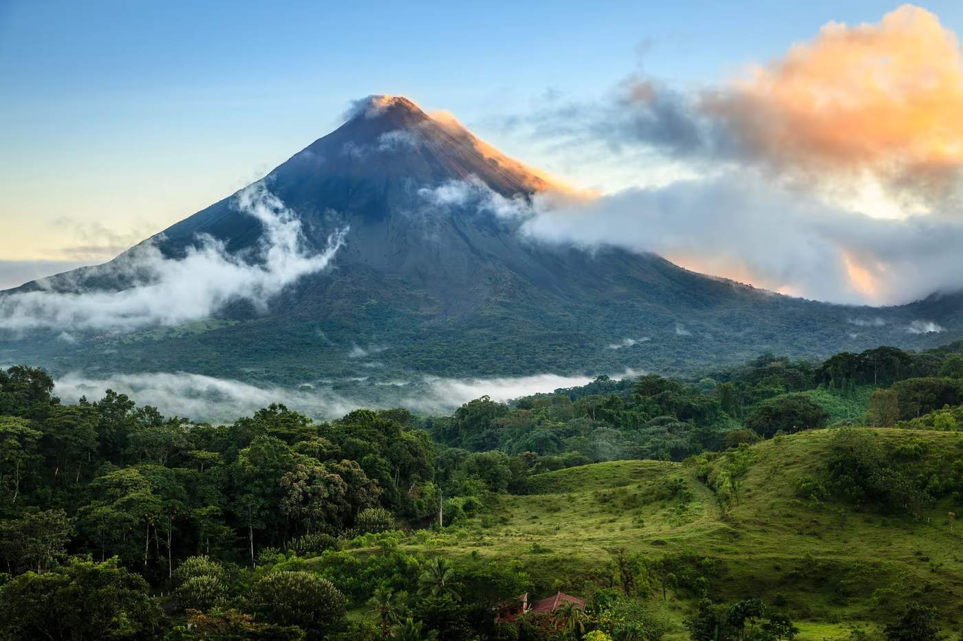 Vue du volcan Arenal au Costa Rica avec des nuages couvrant partiellement le sommet et une végétation luxuriante au premier plan.