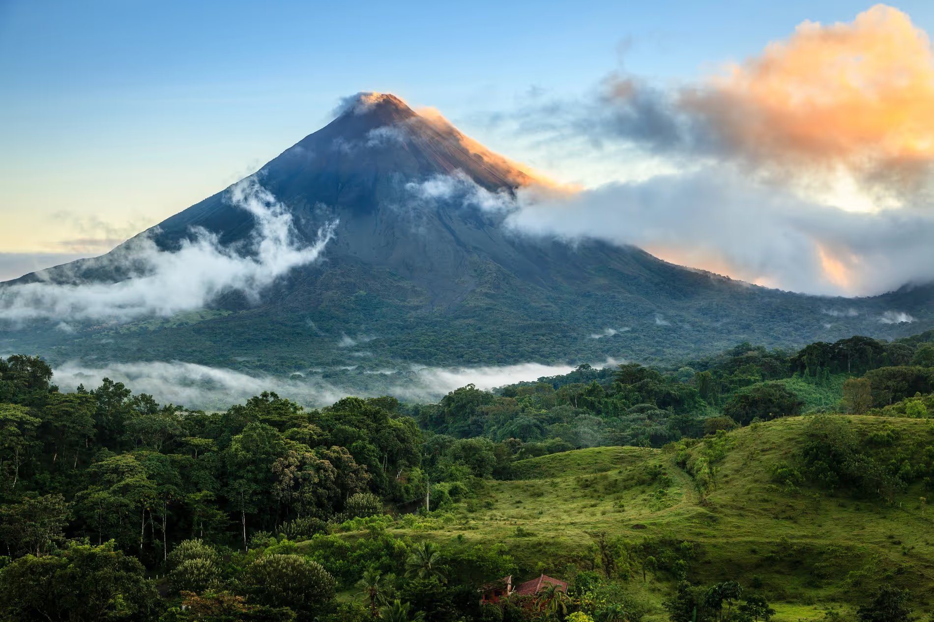 Vue du volcan Arenal au Costa Rica avec des nuages couvrant partiellement le sommet et une végétation luxuriante au premier plan.