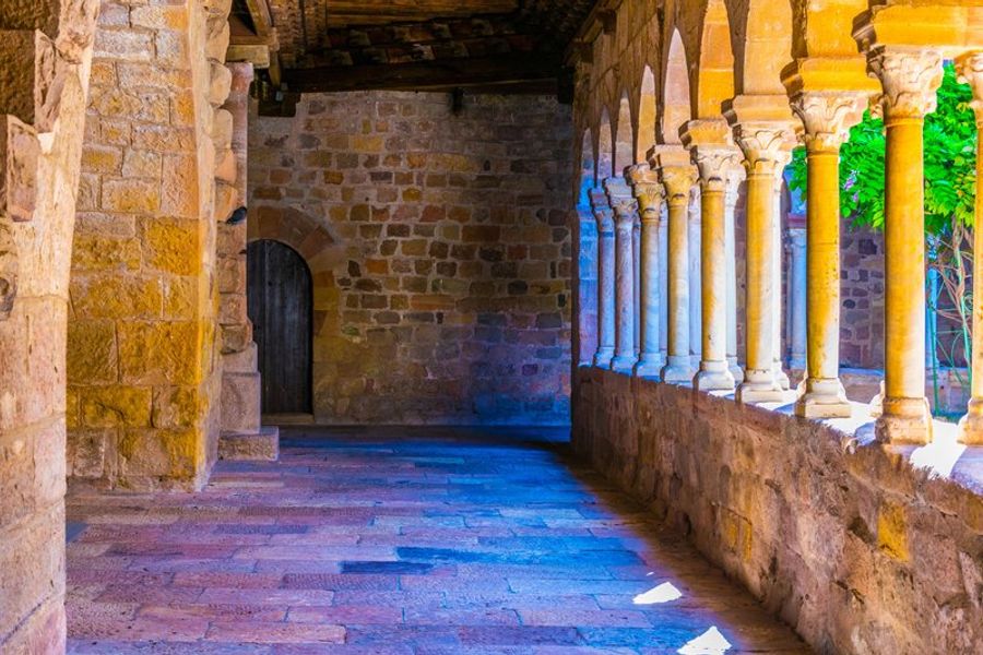 Galerie de cloître historique avec des colonnes sculptées en pierre, baignée de lumière douce et donnant sur une cour verdoyante.