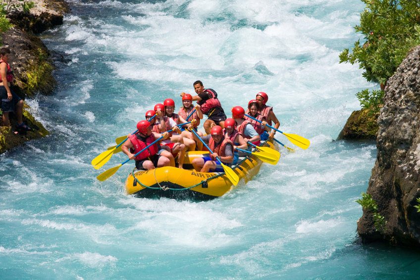Équipe en rafting sur une eau turquoise agitée, portant casques rouges et gilets de sauvetage, dans un canot gonflable jaune encadré de rochers.