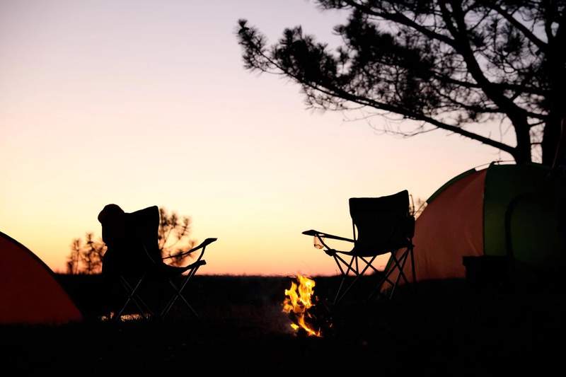 Campement au coucher du soleil avec deux chaises de camping et un feu allumé devant une tente.