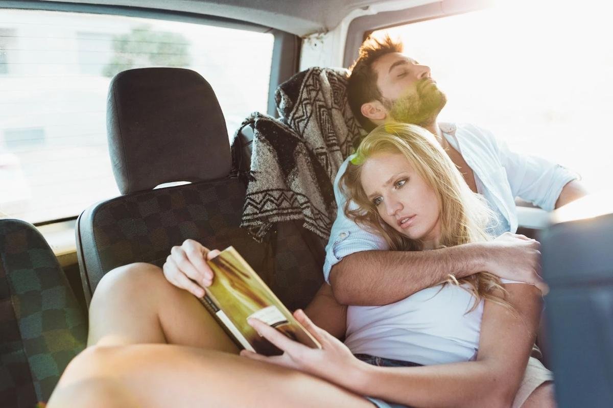 Un couple assis à l’arrière d’une voiture, la femme lisant un livre tandis que l’homme repose sa tête en arrière, profitant d’un moment de détente.