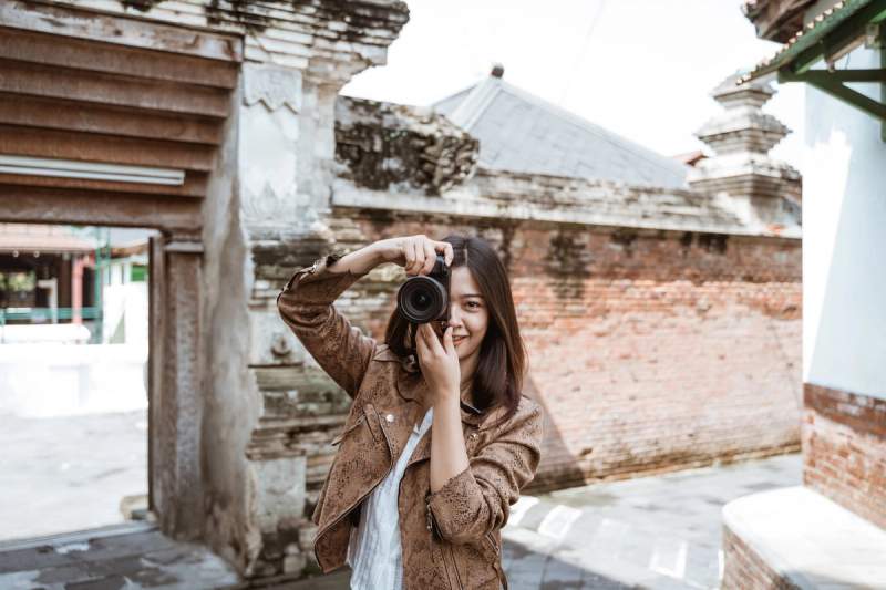 Une jeune femme souriante, vêtue d'une veste marron, prend une photo avec son appareil photo reflex devant une architecture ancienne en briques rouges.