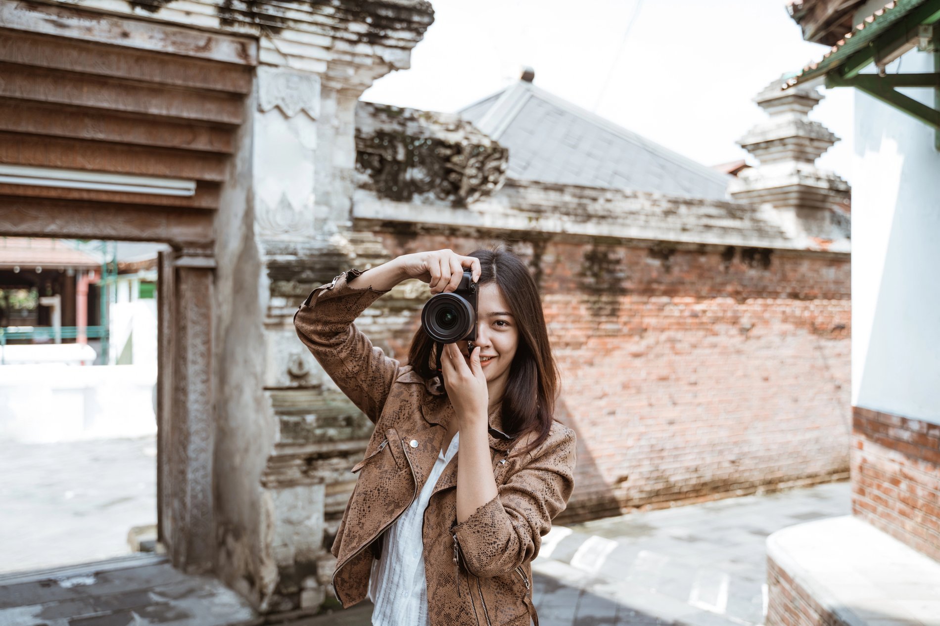 Une jeune femme souriante, vêtue d'une veste marron, prend une photo avec son appareil photo reflex devant une architecture ancienne en briques rouges.