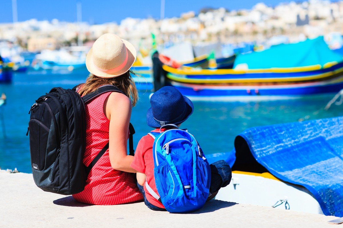 Voyage en famille avec enfant assis face au port, sacs à dos et bateaux colorés en arrière-plan