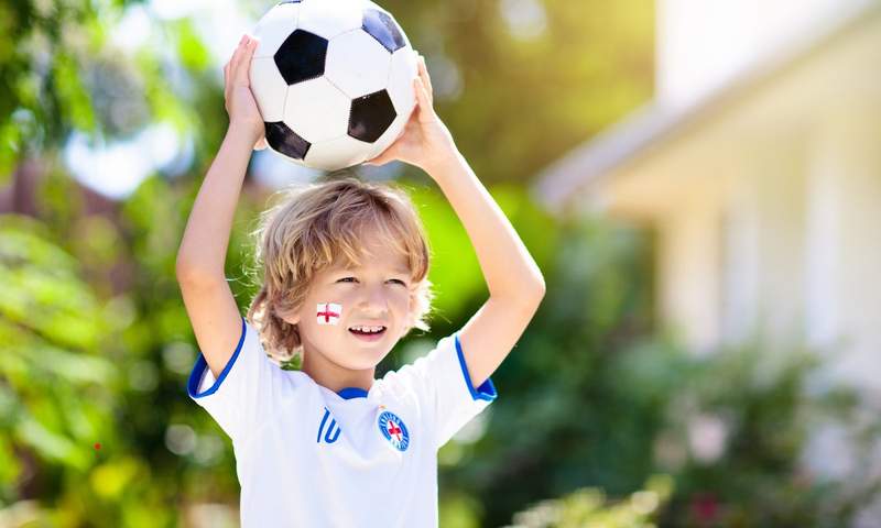 Jeune garçon avec un maillot de football et un ballon, souriant à l’extérieur, avec un drapeau anglais peint sur la joue, symbolisant un road trip en famille sur les traces du football en Angleterre.