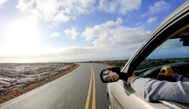 Bras passager posé sur la portière d'une voiture en road trip sur une route panoramique bordée de champs de lave, sous un ciel bleu nuageux.