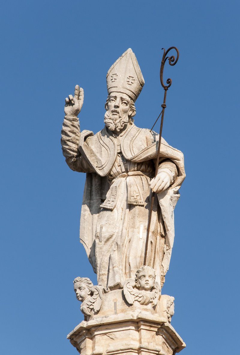 Statue de Sant’Oronzo, représentant un évêque en pierre sculptée avec une crosse, surplombant une place en Italie sous un ciel bleu.