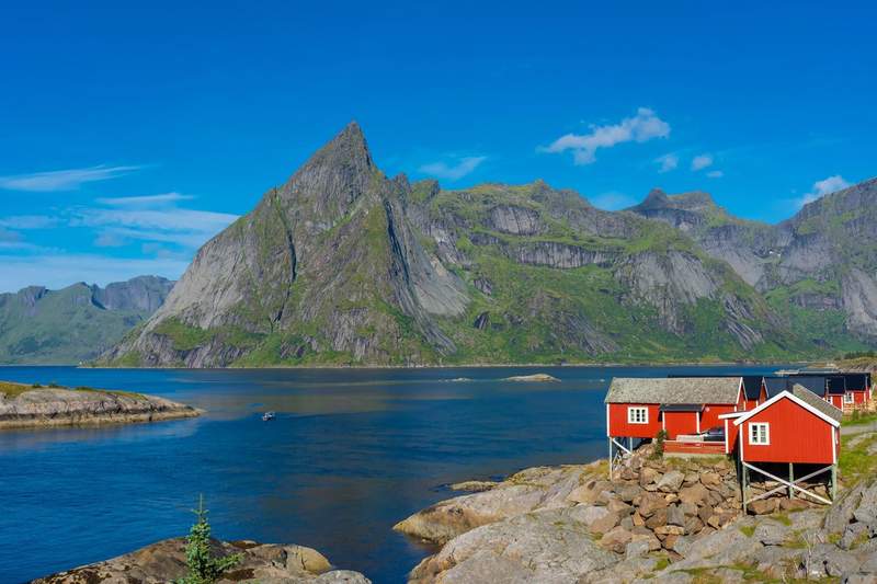Cabanes rouges en bois au bord d’un fjord dans les îles Lofoten, Norvège, avec montagnes verdoyantes en arrière-plan.