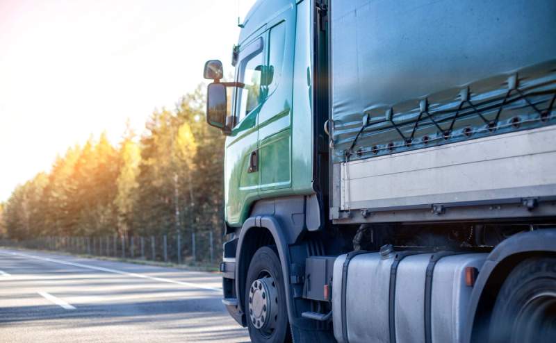 Vue latérale d'un camion avec une semi-remorque verte roulant sur une route bordée de forêts sous un soleil éclatant.