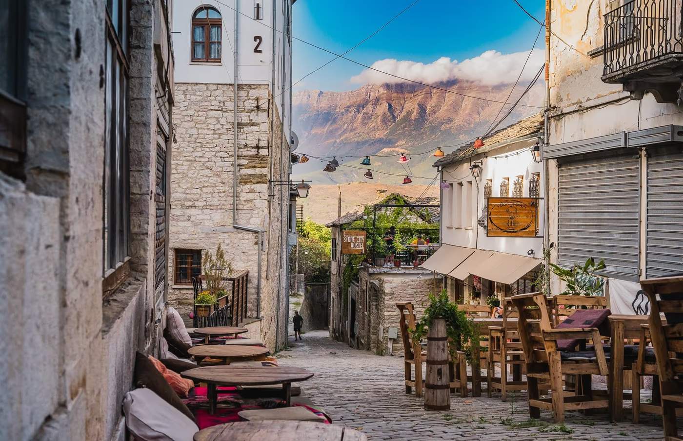 Ruelle pavée de Gjirokastër, Albanie, avec des tables en terrasse et vue sur les montagnes en arrière-plan.