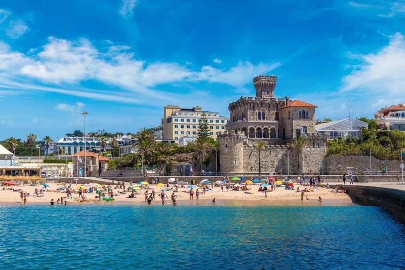 Vue de la plage d'Estoril au Portugal, avec des baigneurs et un château en arrière-plan.