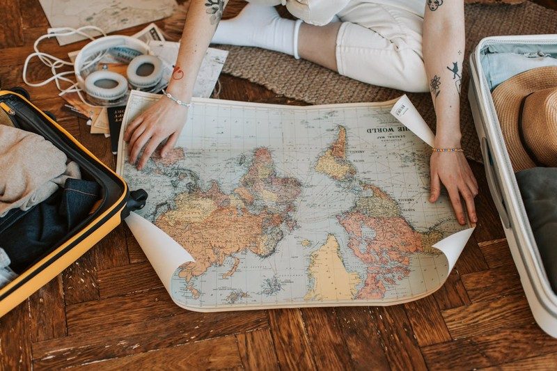 Personne préparant un voyage, examinant une carte du monde entre deux valises ouvertes contenant des vêtements et un chapeau.