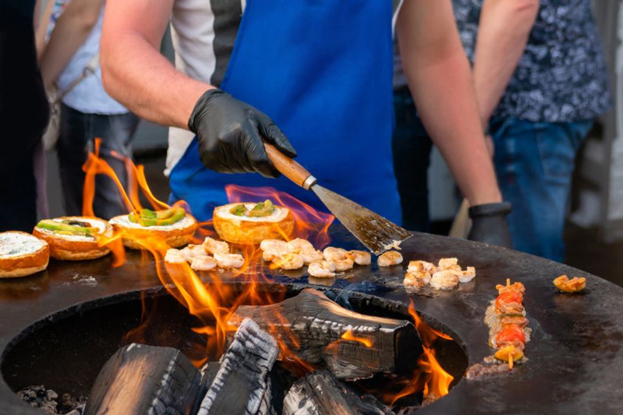 Un cuisinier en plein air préparant des brochettes et des hamburgers sur un barbecue, illustrant la diversité culinaire de la Croatie avec des mets grillés et des spécialités locales.