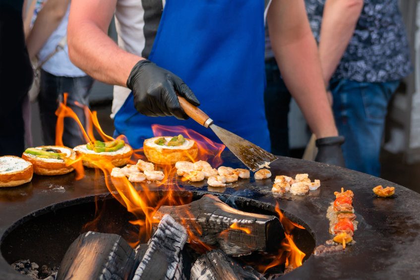 Un cuisinier en plein air préparant des brochettes et des hamburgers sur un barbecue, illustrant la diversité culinaire de la Croatie avec des mets grillés et des spécialités locales.
