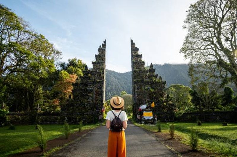 Voyageuse face à la porte traditionnelle d’un temple balinais, entourée de verdure et de montagnes en arrière-plan.