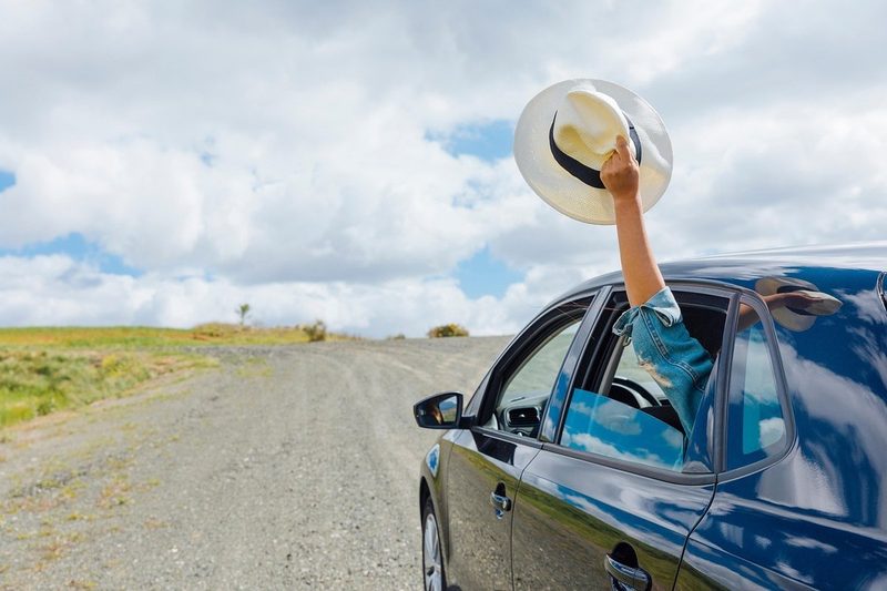 Bras tenant un chapeau en paille sorti par la fenêtre d'une voiture sur une route de gravier, ambiance road trip