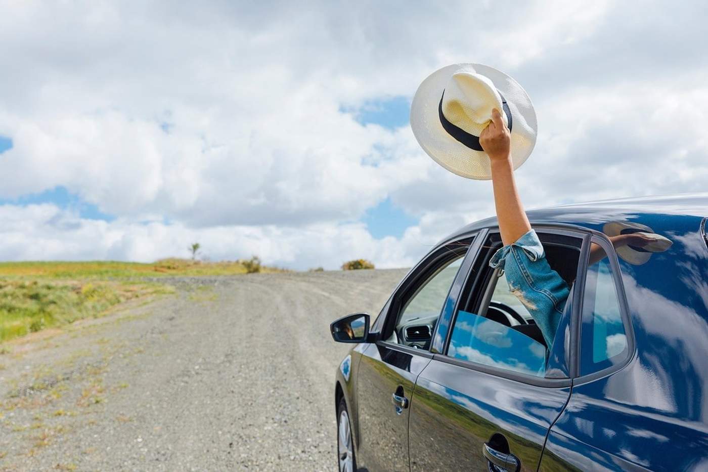 Bras tenant un chapeau en paille sorti par la fenêtre d'une voiture sur une route de gravier, ambiance road trip