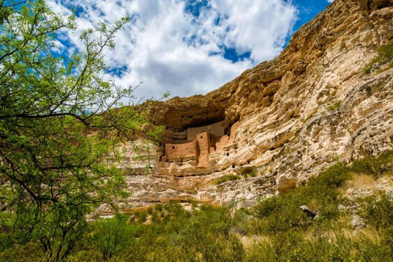 Vue du Château de Montezuma, une habitation troglodytique nichée dans les falaises calcaires de l'Arizona, entourée de verdure et sous un ciel partiellement nuageux.