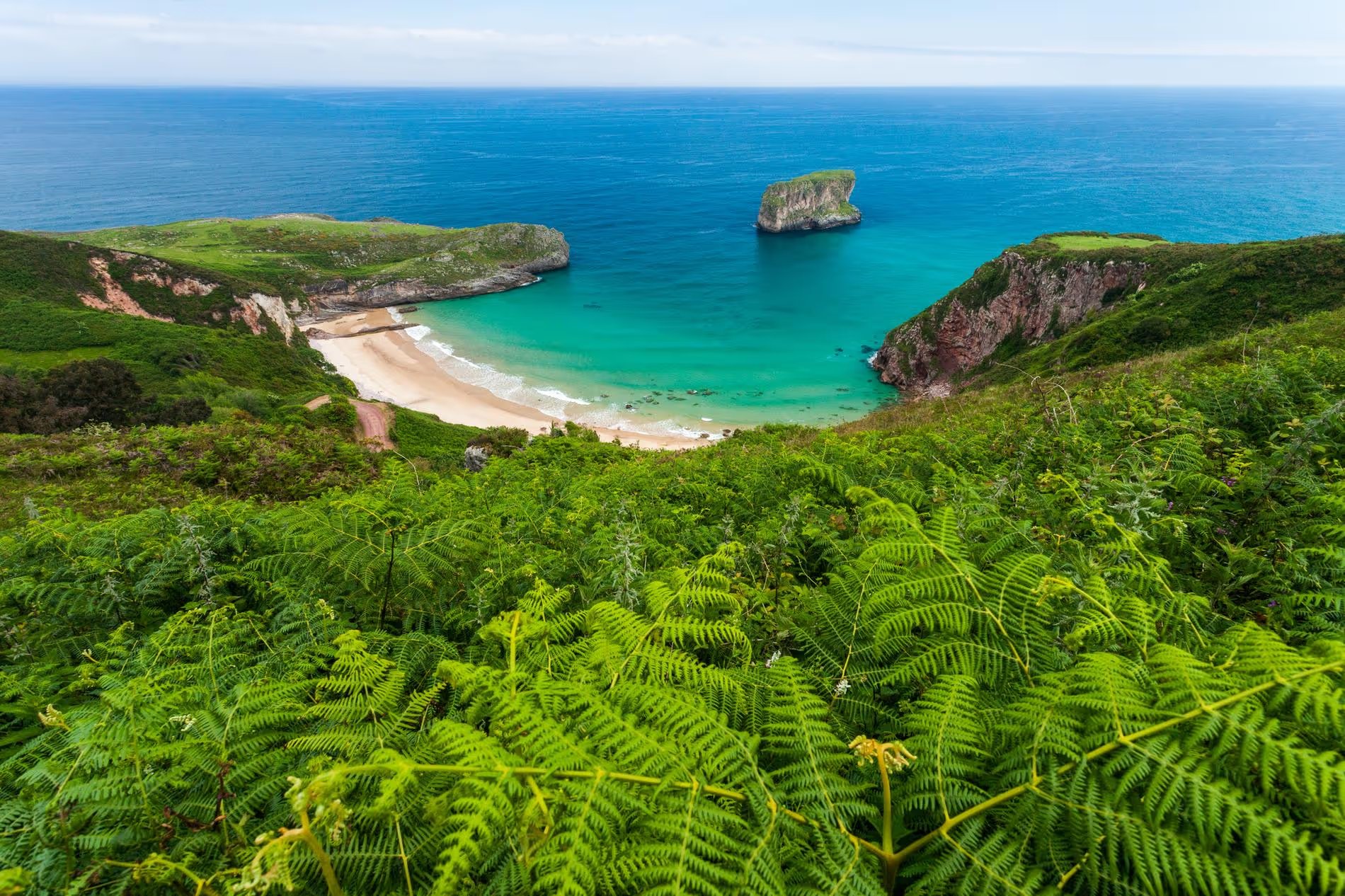 Vue panoramique d’une plage isolée à Llanes entourée de falaises verdoyantes avec une mer bleu turquoise en Espagne.
