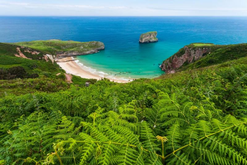 Vue panoramique d’une plage isolée à Llanes entourée de falaises verdoyantes avec une mer bleu turquoise en Espagne.