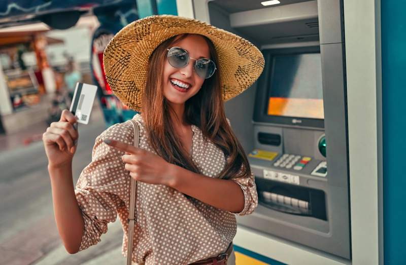 Femme souriante avec un chapeau de paille, tenant une carte bancaire devant un distributeur automatique, illustrant l'utilisation d'une néobanque en voyage.
