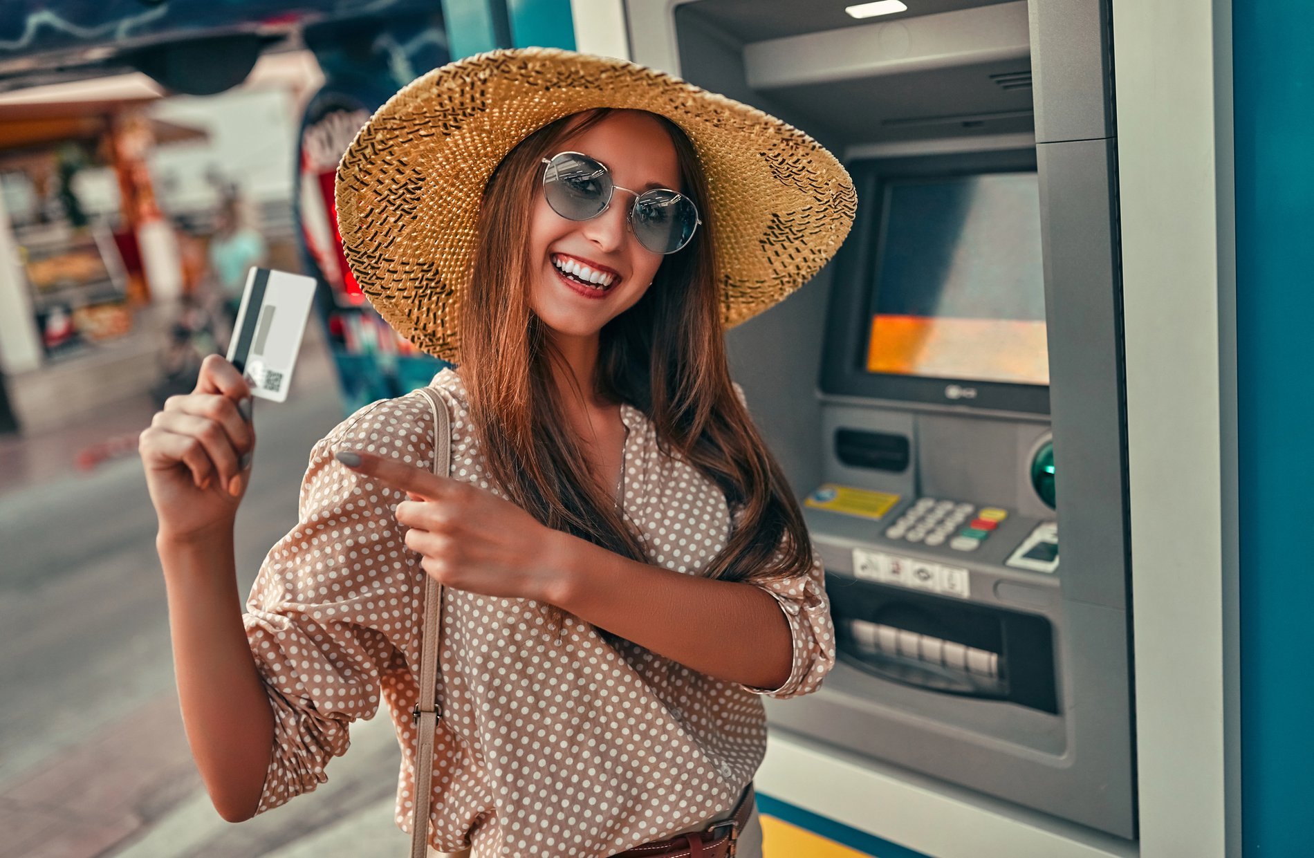 Femme souriante avec un chapeau de paille, tenant une carte bancaire devant un distributeur automatique, illustrant l'utilisation d'une néobanque en voyage.