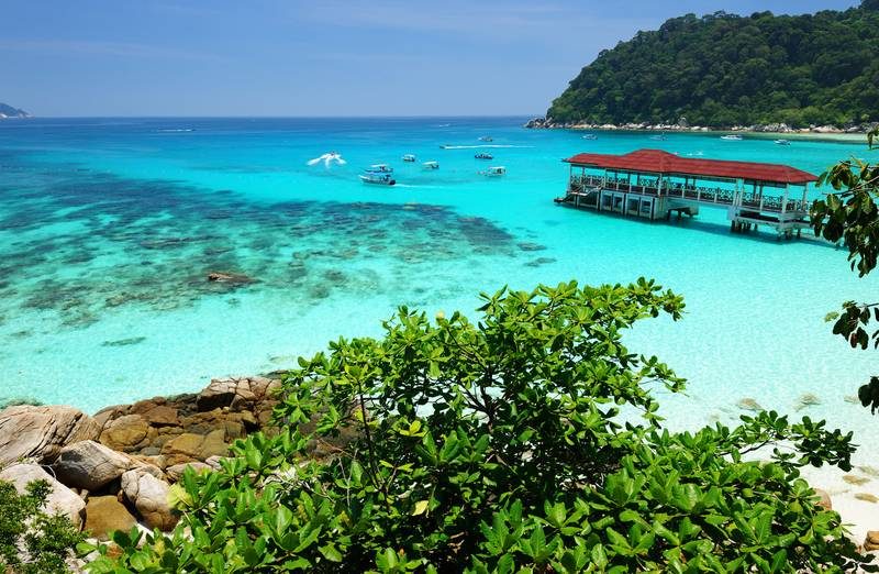 Eau turquoise cristalline bordée de verdure tropicale, bateaux de plaisance et ponton en bois sur une plage en Malaisie.