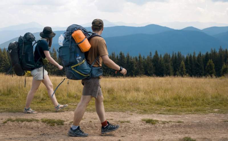 Deux randonneurs équipés de sacs à dos marchant sur un sentier en montagne avec vue sur les forêts et les sommets au loin.