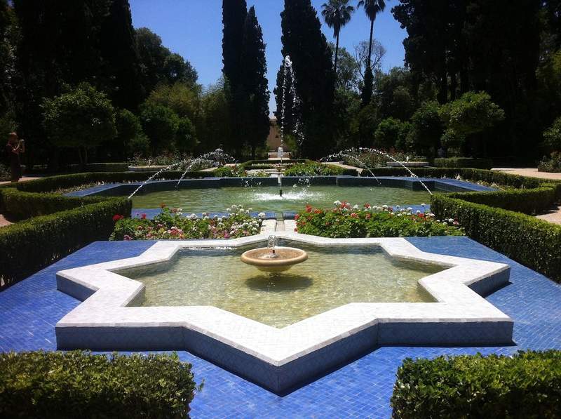 Fontaine décorative en étoile au cœur d’un jardin andalou, entourée de haies fleuries et de jets d’eau symétriques sous un ciel bleu.