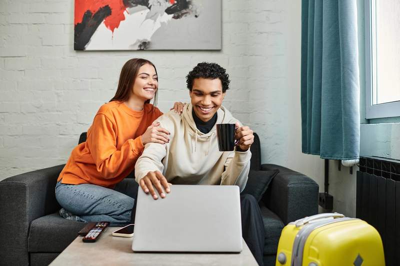 Couple souriant assis sur un canapé, regardant un ordinateur portable pour planifier un voyage, avec une valise jaune au premier plan