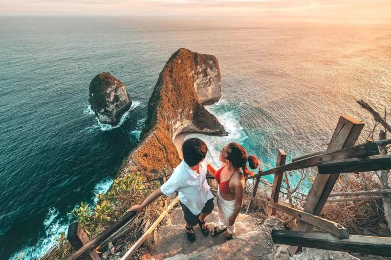 Vue panoramique d’un couple admirant la plage isolée et les formations rocheuses de l’île de Nusa Penida, au coucher du soleil, avec une mer bleue intense en arrière-plan.