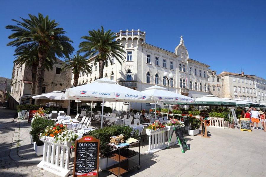 Terrasse animée de restaurants dans la vieille ville de Trogir, Croatie, avec palmiers et architecture historique.