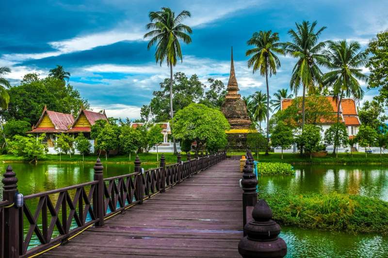 Temple bouddhiste entouré de palmiers en Thaïlande, accessible par une passerelle en bois sur l'eau