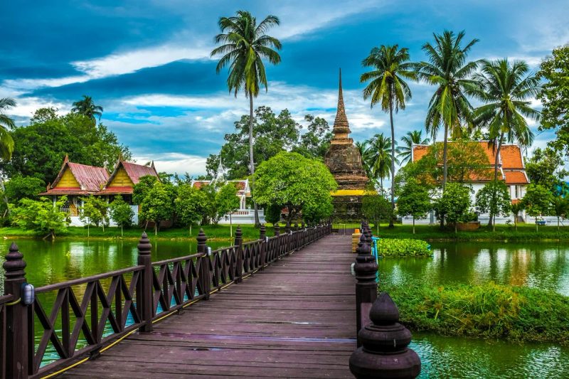 Temple bouddhiste entouré de palmiers en Thaïlande, accessible par une passerelle en bois sur l'eau