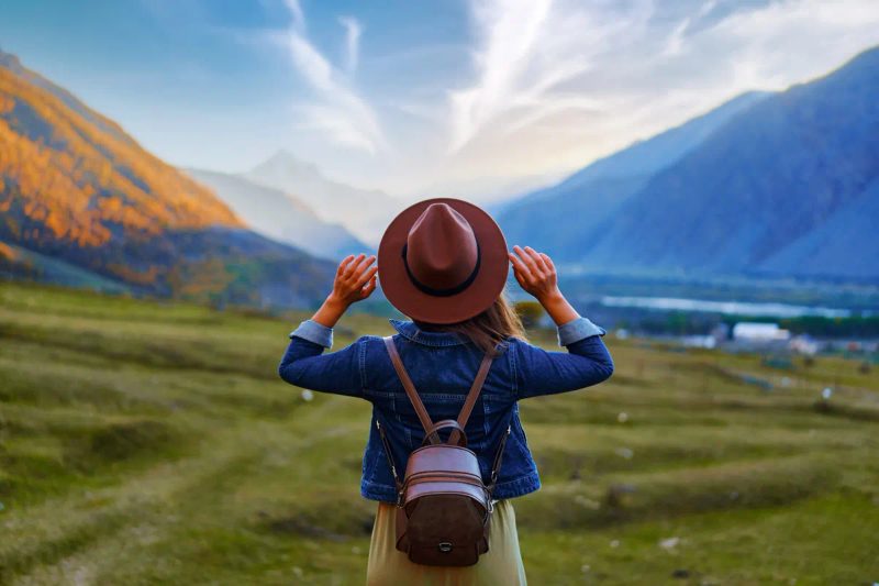 Voyageuse de dos avec chapeau et sac à dos contemplant un panorama de vallée montagneuse aux couleurs d'automne, esprit d'aventure et liberté