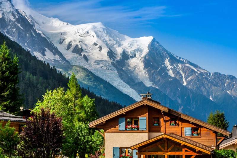 Chalet en bois typique de Chamonix avec des volets bleus et des fleurs aux fenêtres, au pied du Mont Blanc sous un ciel bleu clair.